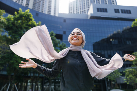 Woman in hijab smiles with arms outstretched in city.