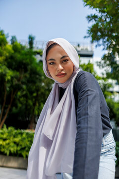 Woman in hijab poses outdoors with greenery backdrop.