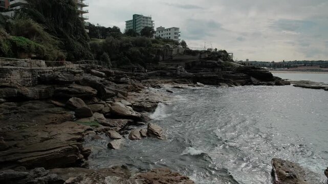 Scenic rocky coastline along the famous Bondi to Bronte Coastal Walk in Sydney. Waves crash against the sandstone shore near Tamarama with apartments on the cliff.