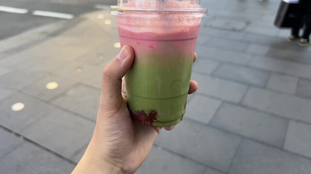 POV of hand holding a layered strawberry matcha latte in a plastic takeaway cup. Iced pink and green tea beverage with a straw, held over a city sidewalk. Refreshing summer drink.