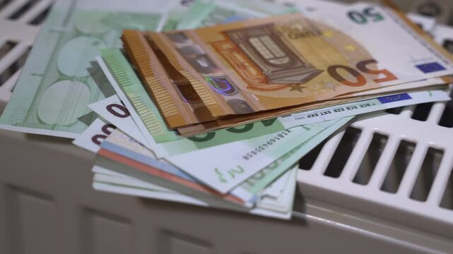 Pile of mixed euro banknotes on a warm radiator, symbolizing the financial burden of heating costs, rising energy prices and increasing winter household expenses across Europe.