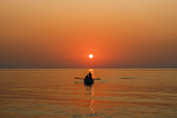 Silhouettes of people in a boat rowing across calm water against a bright orange sea sunset