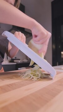 Close-up of Asian woman shredding cabbage using a mandoline slicer on a wooden cutting board in a home kitchen, vertical format