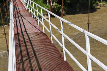 Long metal suspension bridge with red path and white railing offers pedestrian crossing over muddy...