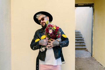 A bearded man in a hat holds flowers and disposable cups.
