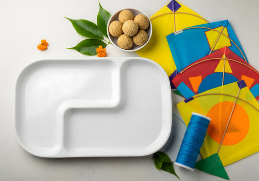 Makar Sankranti festival background with empty food plate, colourful kites, manja spool, tilgul ladoo, leaves and decorations arranged on a clean surface for celebration and festive presentations