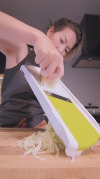 Close-up of Asian woman shredding cabbage using a mandoline slicer on a wooden cutting board in a home kitchen, vertical format