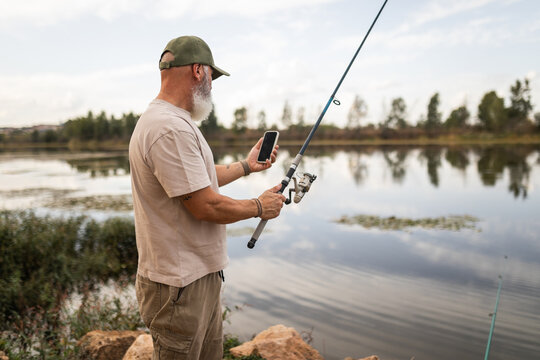 Mature bearded man fishing with phone by the river