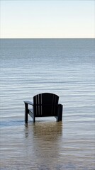 A black chair stands in the water, with a vast ocean and sky in the background, creating a serene and isolated scene.