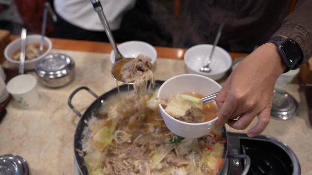 Hand Serving Traditional Korean Hot Pot (Jeongol) with Meat and Noodles into Bowl, Close-up of Ladle Scooping Bulgogi and Vegetables from Boiling Asian Stew