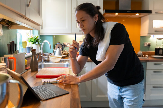 Thoughtful Man Writes Grocery List in Kitchen While Checking Tablet