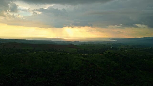 A rising sunset aerial capturing golden rays breaking through clouds over rolling green hills and distant valleys, illuminating the serene landscape in Mariveles, Bataan, Philippines