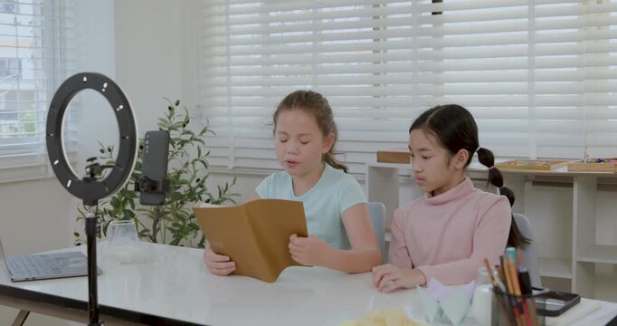kids creating content for social media. A Caucasian girl reads from a book while recording a video on a smartphone with a ring light, accompanied by her Asian friend. Young vlogger storytelling 4k 