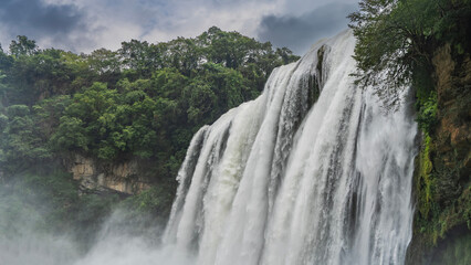 Obraz premium A picturesque tropical waterfall. Streams of white water flow down from a sheer cliff. Foam, splashes. Green vegetation. Clouds in the sky. China. Huangguoshu Waterfall. Guizhou
