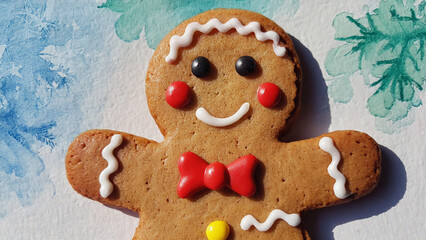 Gingerbread man cookie with icing and red bow tie on a sheet of paper decorated with watercolor snowflakes and paint spots, with colored pencils nearby. Concept of Christmas creativity and kids winter