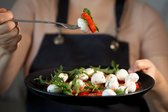 A person in a black apron enjoys a Caprese salad in the kitchen during the day. A fork holds a mozzarella ball, bell pepper, and leaf near the plate.
