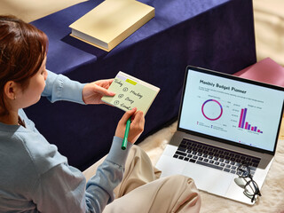 A woman plans her day by taking notes while using a laptop