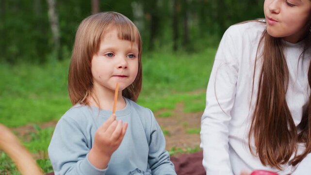 little girl sitting on picnic mat beside sister eating breadsticks and smiling cheerfully while sister wipes leftover from lips surrounded by picnic snacks basket and green background