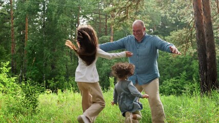 Playful dad spreading hands wide with joyful smile waiting for daughters running to hug him during happy family hangout in lush green forest under warm sunlight surrounded by tall trees and grass