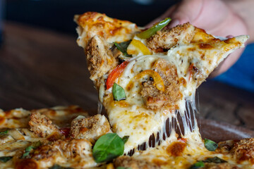 Close-up of a cheesy pizza slice loaded with fried chicken, peppers, basil, and melted mozzarella...