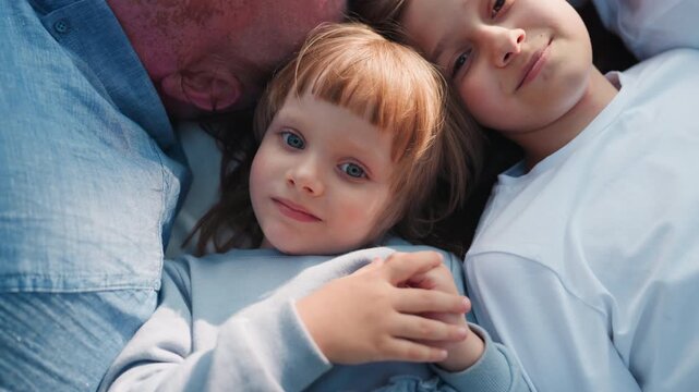 Zoom in on little damsel lying between sister and dad during relaxing outdoor moment, smiling softly under warm sunlight, capturing peaceful childhood bond and calm family connection