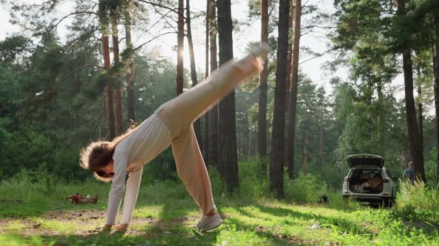 Youth practicing cartwheel on picnic mat in sunlit forest clearing near family car, surrounded by trees and soft sunlight, showing energy, joy, and carefree movement during peaceful summer outdoor