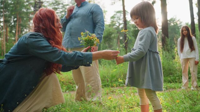 Mother hands flower to daughter to smell during peaceful walk in green forest, surrounded by sunlight and nature, capturing gentle moment of love, tenderness, and connection in family outdoor life