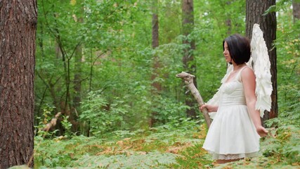 Female goddess with white wings walks through lush woodland holding wooden staff, surrounded by green ferns and towering trees, radiating grace, strength, purity, and mystical presence