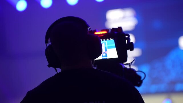 Event videographer filming stage performance with professional camera rig, wearing headphones and monitoring live feed in blue illuminated venue environment during production