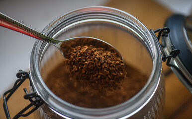 A spoon dipped into a glass jar filled with instant coffee granules on a wooden table, captured in warm natural light.