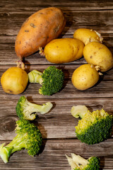 A mix of fresh raw produce, including potatoes, a sweet potato and broccoli florets sit on a rustic wooden surface, ready to be cooked.