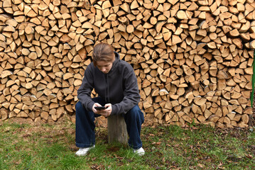 Young woman sitting on a tree stump using a smartphone in front of a large firewood stack