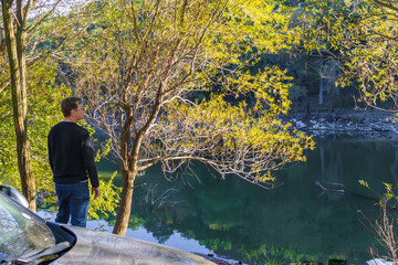 A traveler stands by a parked car in a forested area, enjoying a quiet outdoor moment during a road and hiking trip.