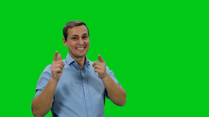Young smiling man in light blue colored shirt pointing fingers at you against green background