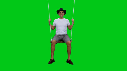 Young smiling man tourist in white T-shirt and black cowboy hat sits on rope swing against green background