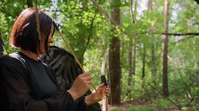 Woman with black wings carefully places arrow on bowstring in green forest, sunlight shining through trees highlighting focus and precision, cinematic moment of concentration