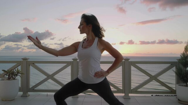 Woman practicing dynamic tai chi poses on a balcony overlooking the ocean during a peaceful morning sunrise.