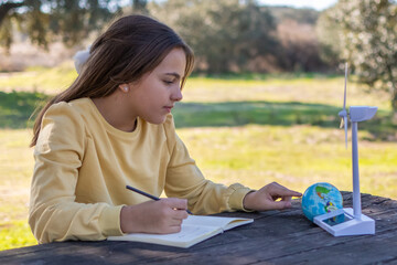 Girl studying environmental science concepts outdoors, writing in a notebook while pointing at a model globe, wind turbine, and solar panel, promoting sustainable education