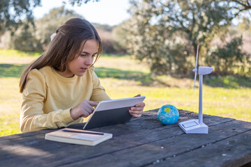 Young girl engaging in outdoor education, using a digital tablet to learn about renewable energy with a mini wind turbine model and a globe on a rustic wooden table