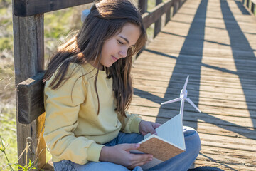 Young girl sits on a wooden bridge studying a notebook with a small wind turbine model, enjoying sunlight, learning about renewable energy and connecting with nature and the future generation