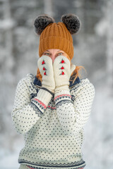Close-up portrait of a young caucasian red hair woman in the winter snowy forest. A young woman Is covering her face with her hands in mittens. Winter leisure in the park.