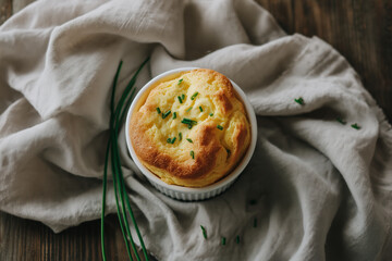 Golden cheese souffle sprinkled with fresh chives, served in a white ramekin over a rustic wooden table with a linen cloth, perfect for brunch concepts