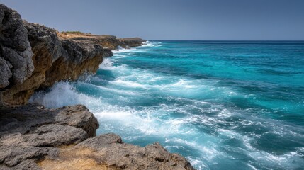Beautiful view of the ocean from a rocky cliff. the sky is clear and blue, and the water is a deep turquoise color. the waves are crashing onto the rocks, creating white foam.