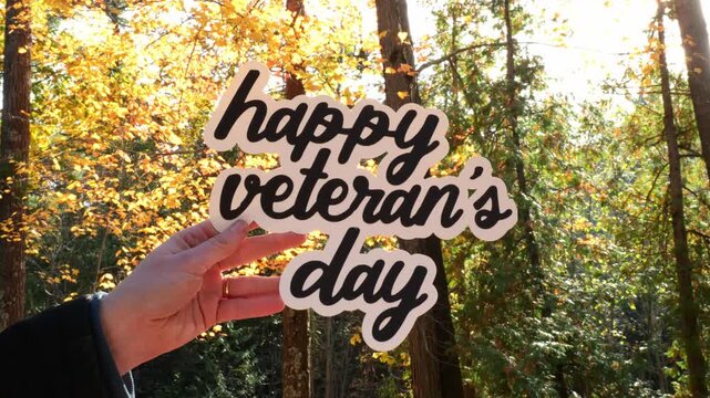 Close Up of Young Woman in Forest Holding Up Words Happy Veteran’s Day in the sunlight With Colorful Fall Leaves 4k Video Stock Footage
