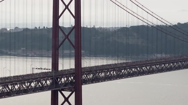 Half marathon and 10K long distance running event in Lisbon, Portugal, Europe. Runners crossing the iconic famous red 25th April suspension bridge. Panning professional aerial drone shot
