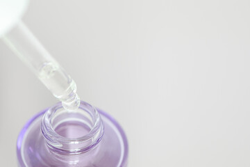 Close-up of a dropper releasing serum into a purple glass bottle on a light background with copy space. Ideal for skincare, cosmetics, beauty products, wellness, and self-care themes.