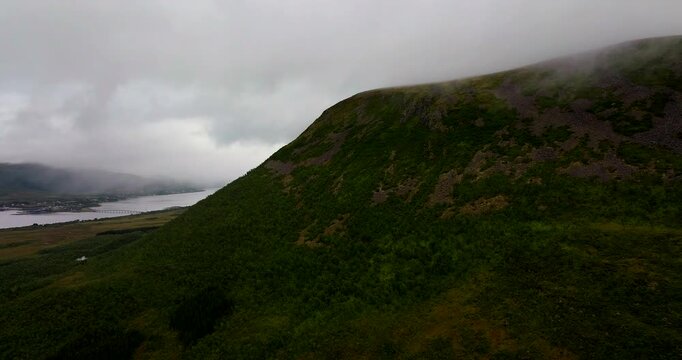 Lush green mountain slope disappearing into low clouds. The town of Sortland and the fjord are visible in the distance under a moody grey sky in Northern Norway. Aerial forward