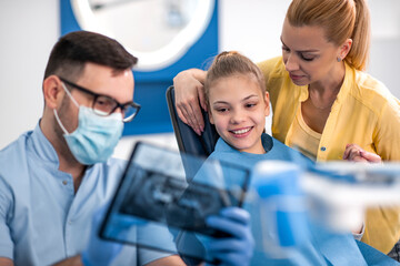 Dentist shows young girl dental X-ray while mother watches in a bright clinic setting