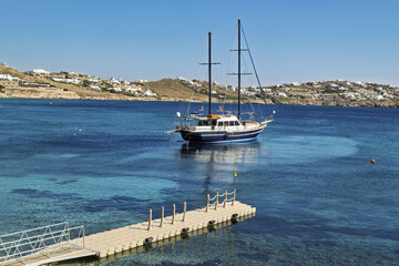 Coastal view in Mykonos, Greece, featuring a large sailboat anchored in clear blue water near a dock. The dock extends from the foreground with railings and posts,