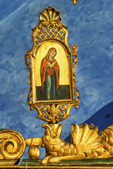 Religious icon in Mykonos, Greece, framed in an ornate golden structure against a blue background. The icon depicts a figure in red and blue robes, likely the Virgin Mary,